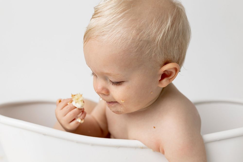 Blonde baby sitting in a white bathtub, holding and examining a small toy or food item.
