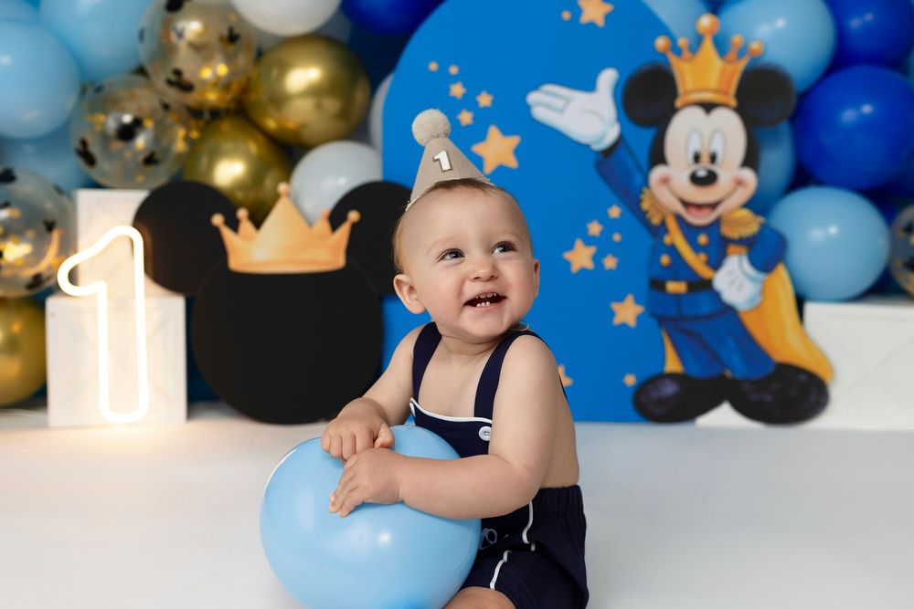 Baby boy in navy romper sits smiling at Mickey Mouse themed first birthday party with blue and gold balloons.