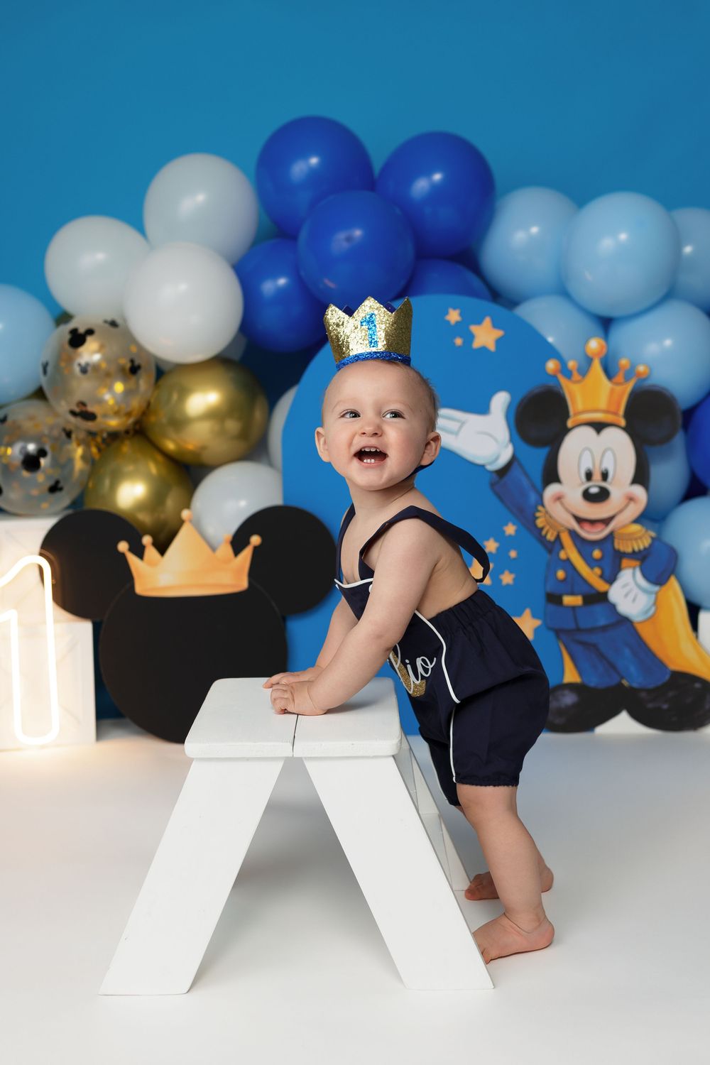 Baby boy in navy romper with crown hat celebrates first birthday with Mickey Mouse balloons backdrop.