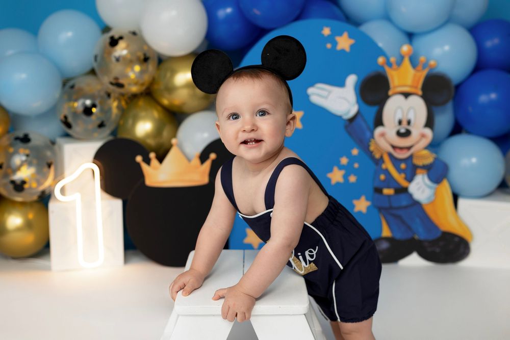 Baby boy in navy romper with Mickey Mouse ears celebrates first birthday with balloon backdrop and Mickey Mouse theme.