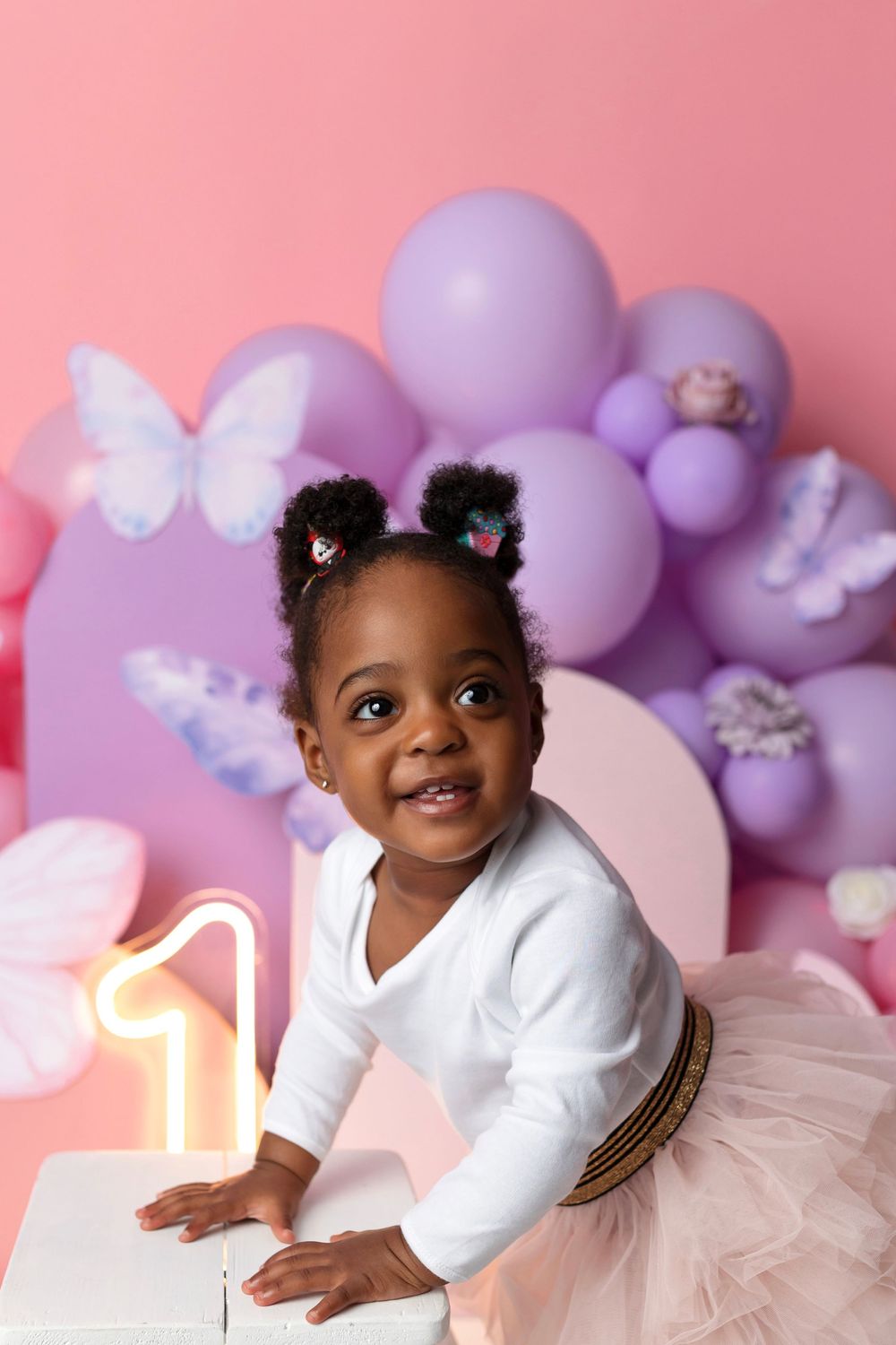 Adorable baby girl in pink tutu celebrates first birthday with purple balloons and butterfly decorations.
