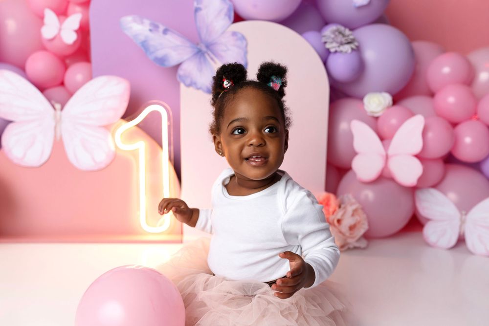 Toddler girl in white top and pink tutu celebrates first birthday with butterfly decorations and colorful balloon backdrop.