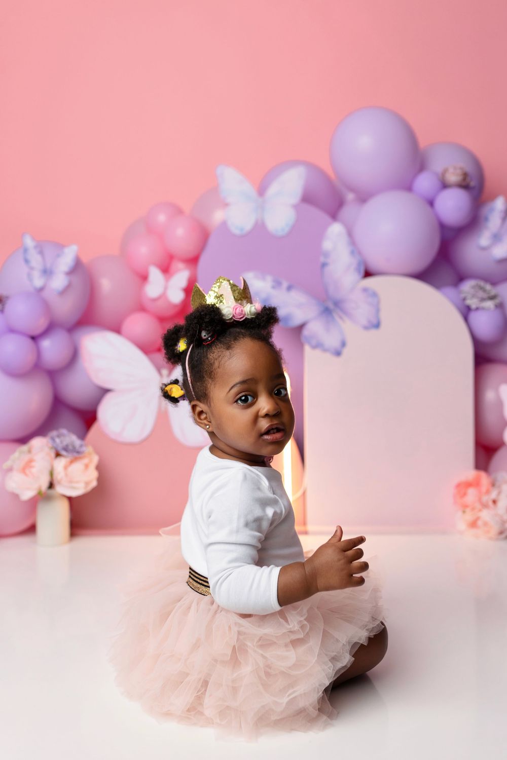 A baby girl in a tutu skirt and gold crown sits before a pink and purple balloon and butterfly birthday backdrop.