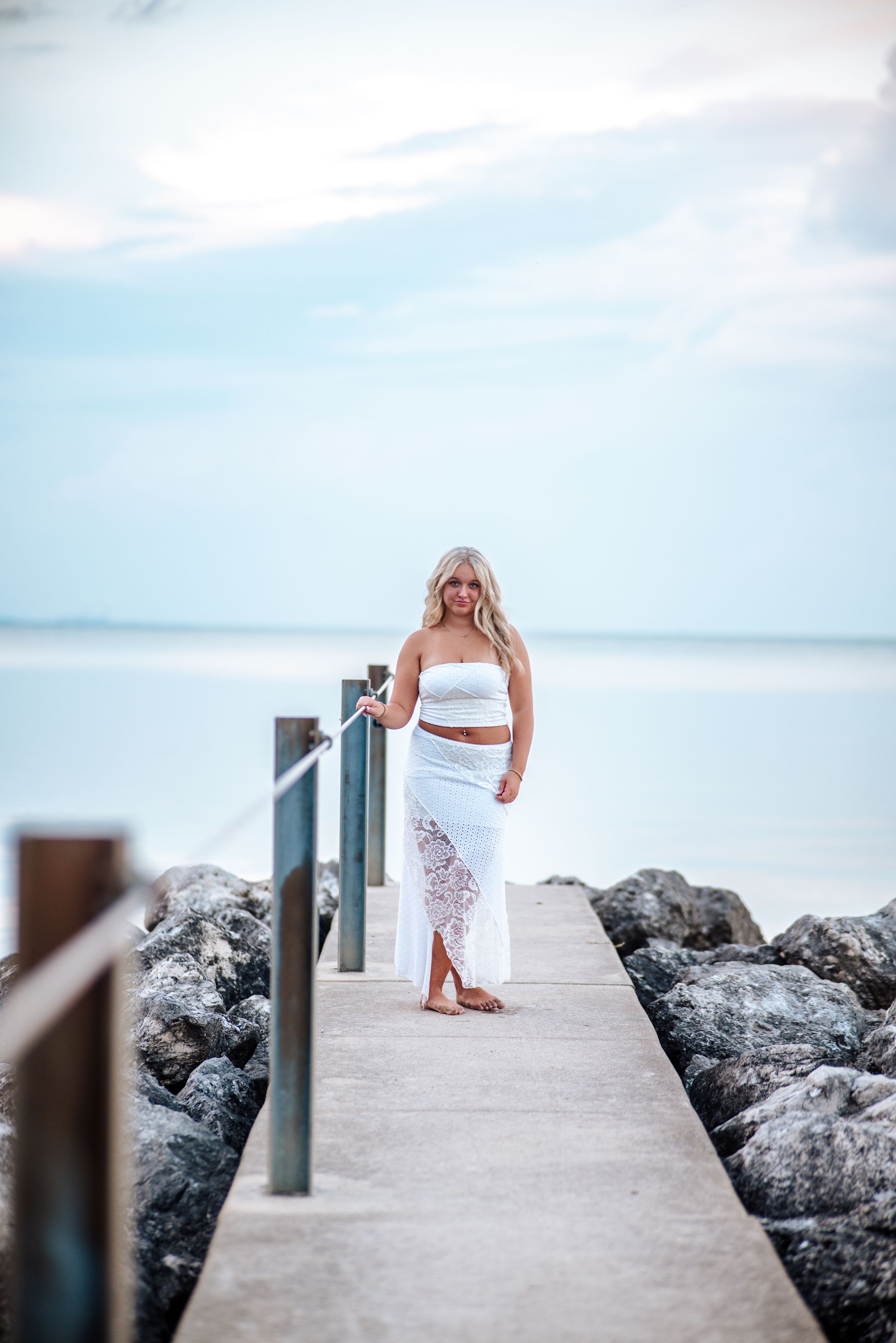A person in a white dress stands on a concrete pier overlooking rocky coastline and ocean on a bright cloudy day.