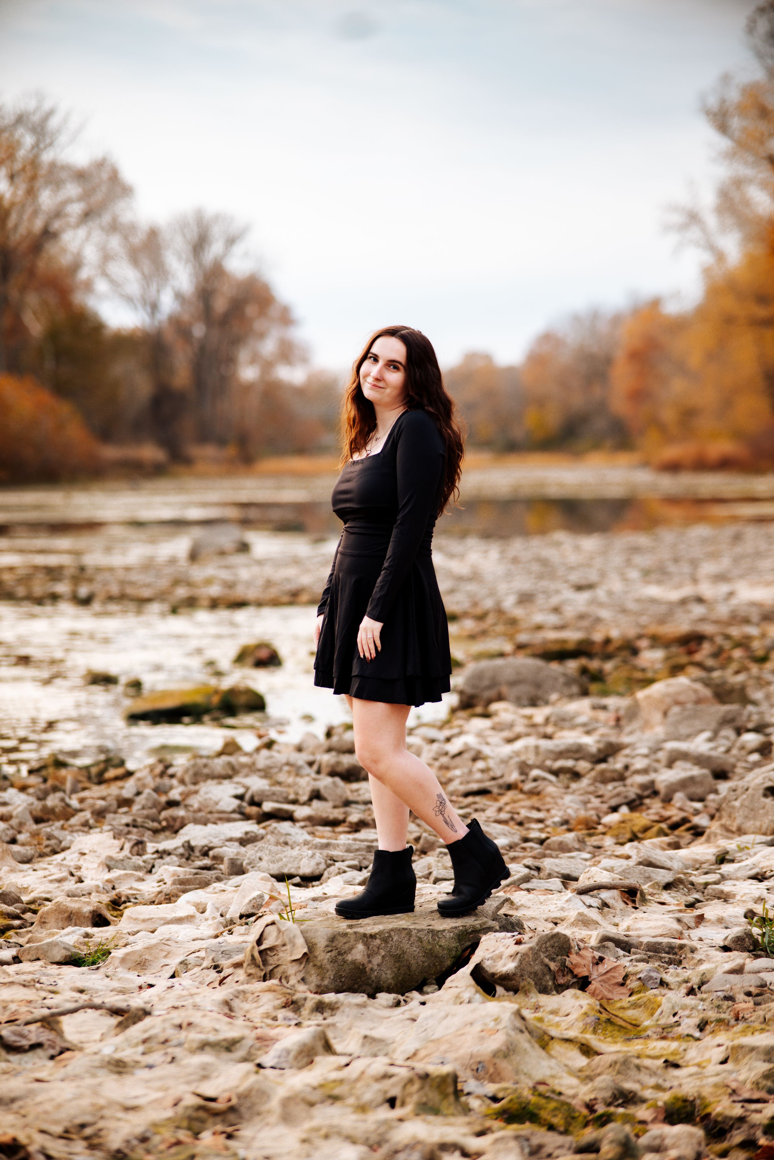 A figure in a black coat poses on rocky terrain during autumn with golden trees in the background.