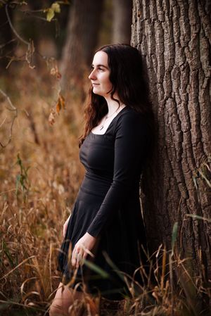 A woman in a black dress poses thoughtfully against a tree trunk surrounded by autumn foliage in a natural outdoor setting.