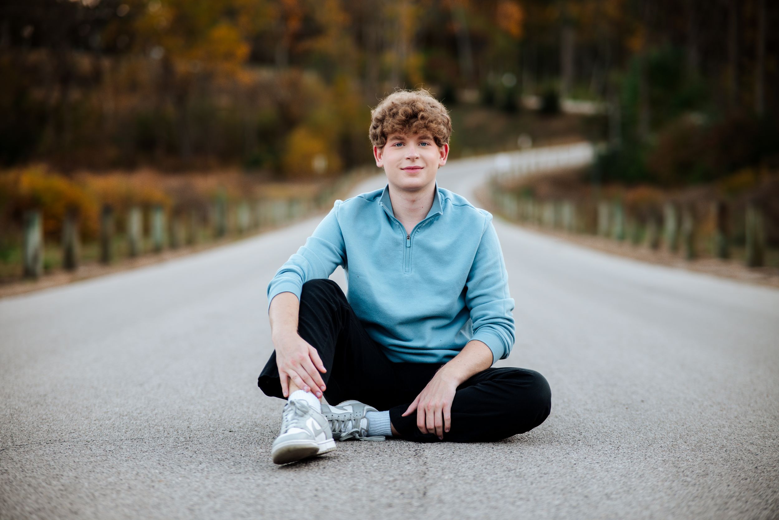 Someone in a light blue sweater sits cross-legged on an empty road during autumn with blurred trees in background.