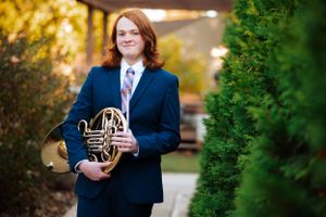 Professional portrait of musician in navy suit holding French horn outdoors.