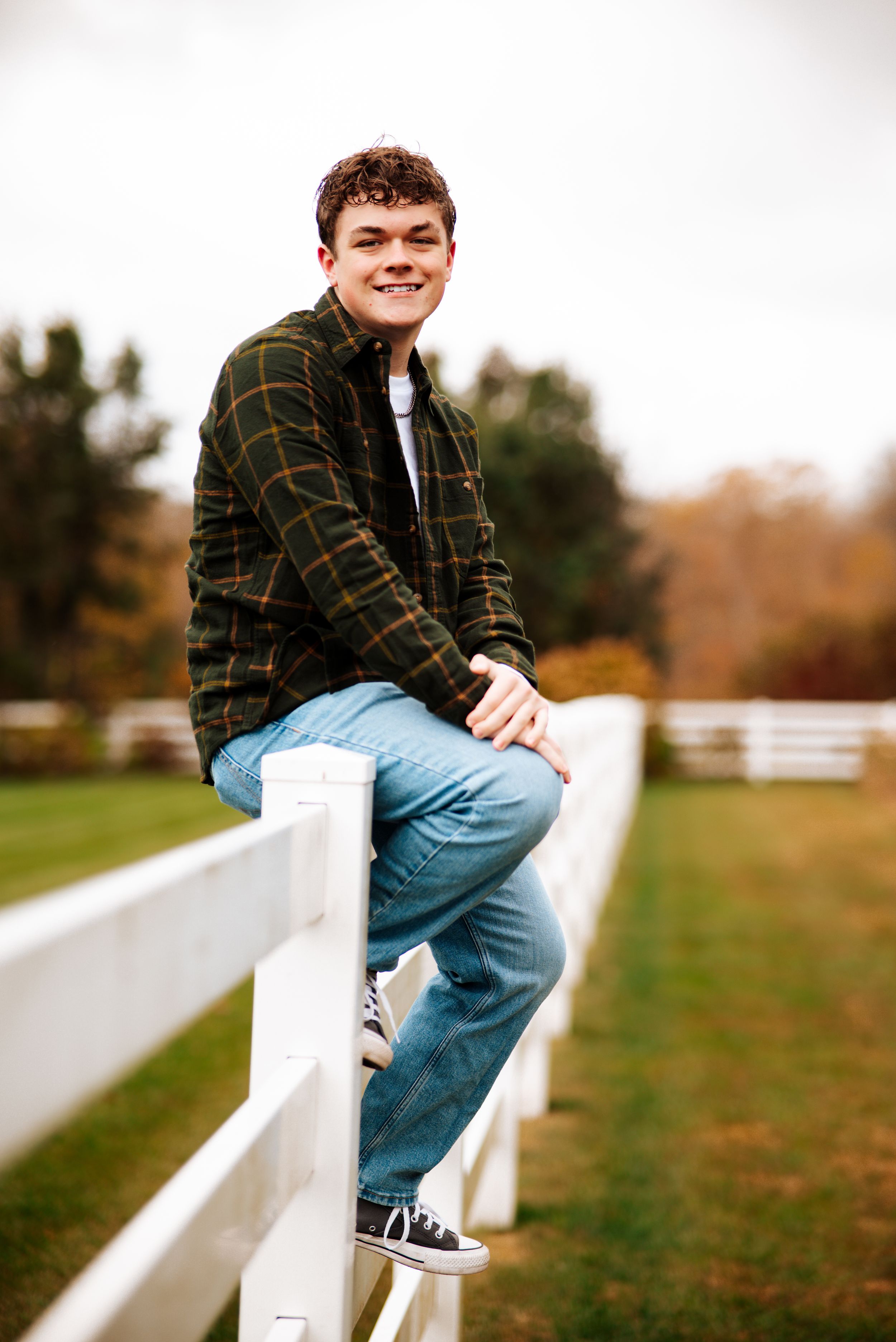 Person in plaid shirt and jeans sits casually on white fence in autumn setting with grass and trees in background.