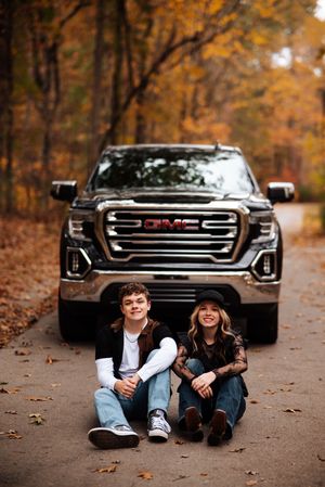 Two people sit and lay on fallen autumn leaves in front of a black GMC truck on a forest road.