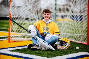 Senior High school lacrosse goalie in blue and yellow uniform makes a diving save during practice on outdoor field in Findlay Ohio