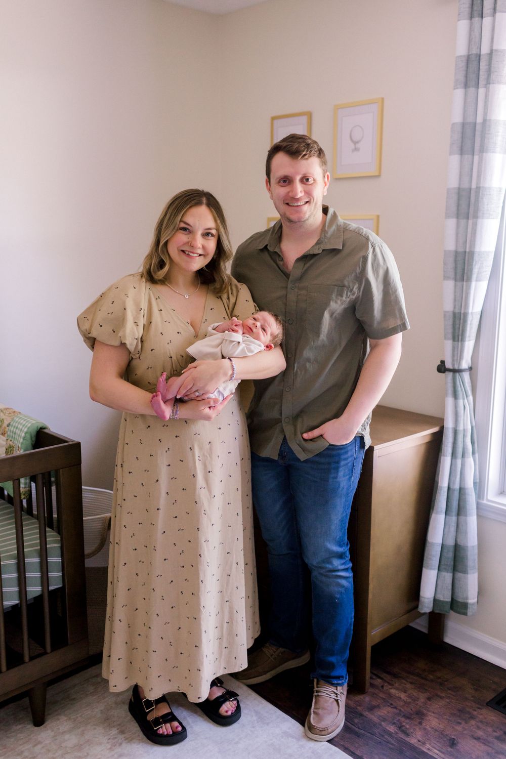 Happy couple standing in nursery holding their newborn baby, mother in floral dress, father in green shirt and jeans.