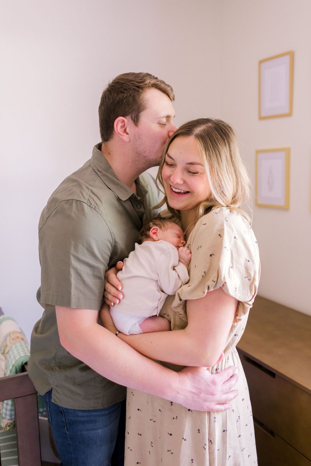 Happy new parents share a tender moment, dad kissing mom on cheek as she smiles holding their newborn baby.