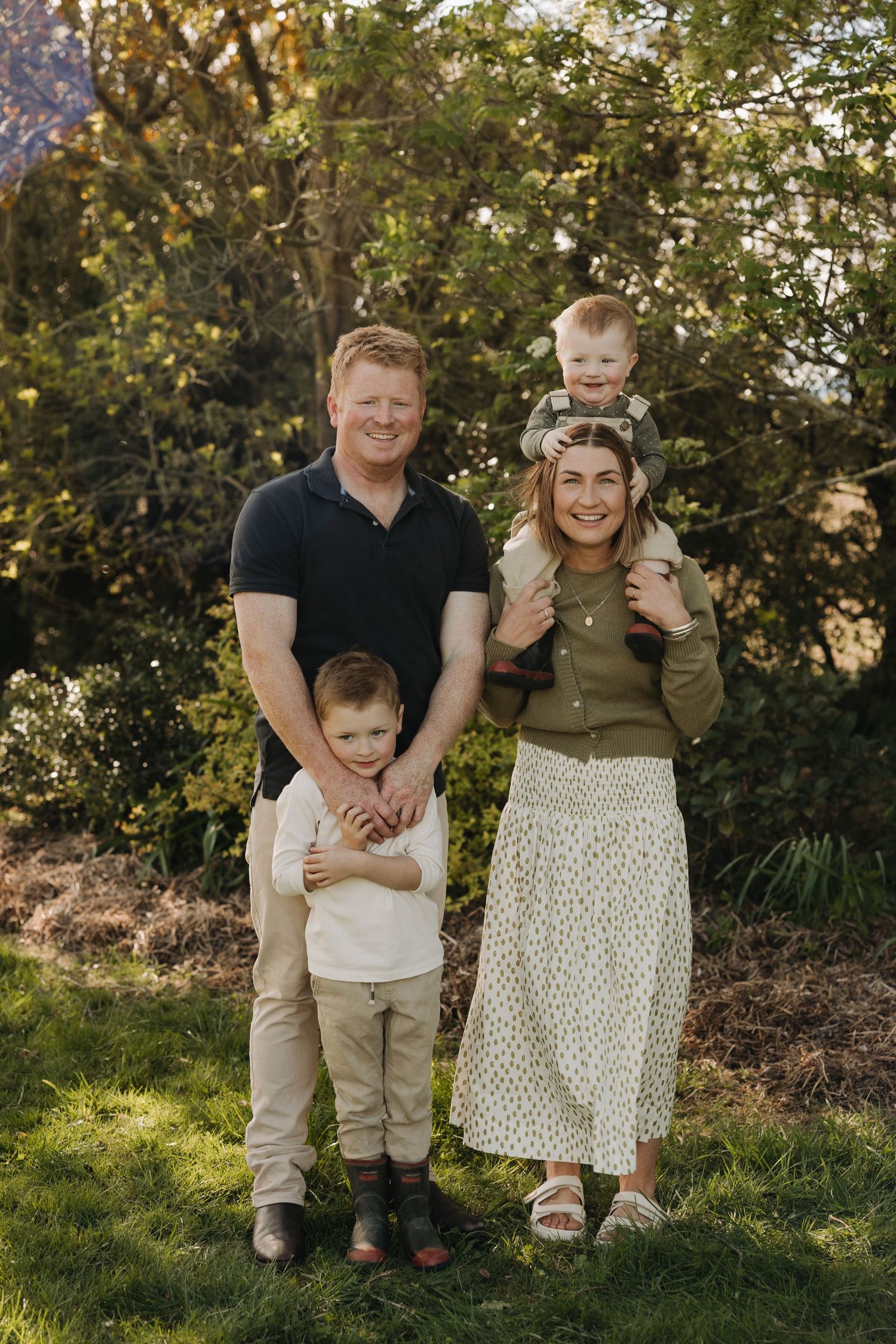 A family poses together outdoors in coordinated neutral outfits against a natural backdrop during golden hour.