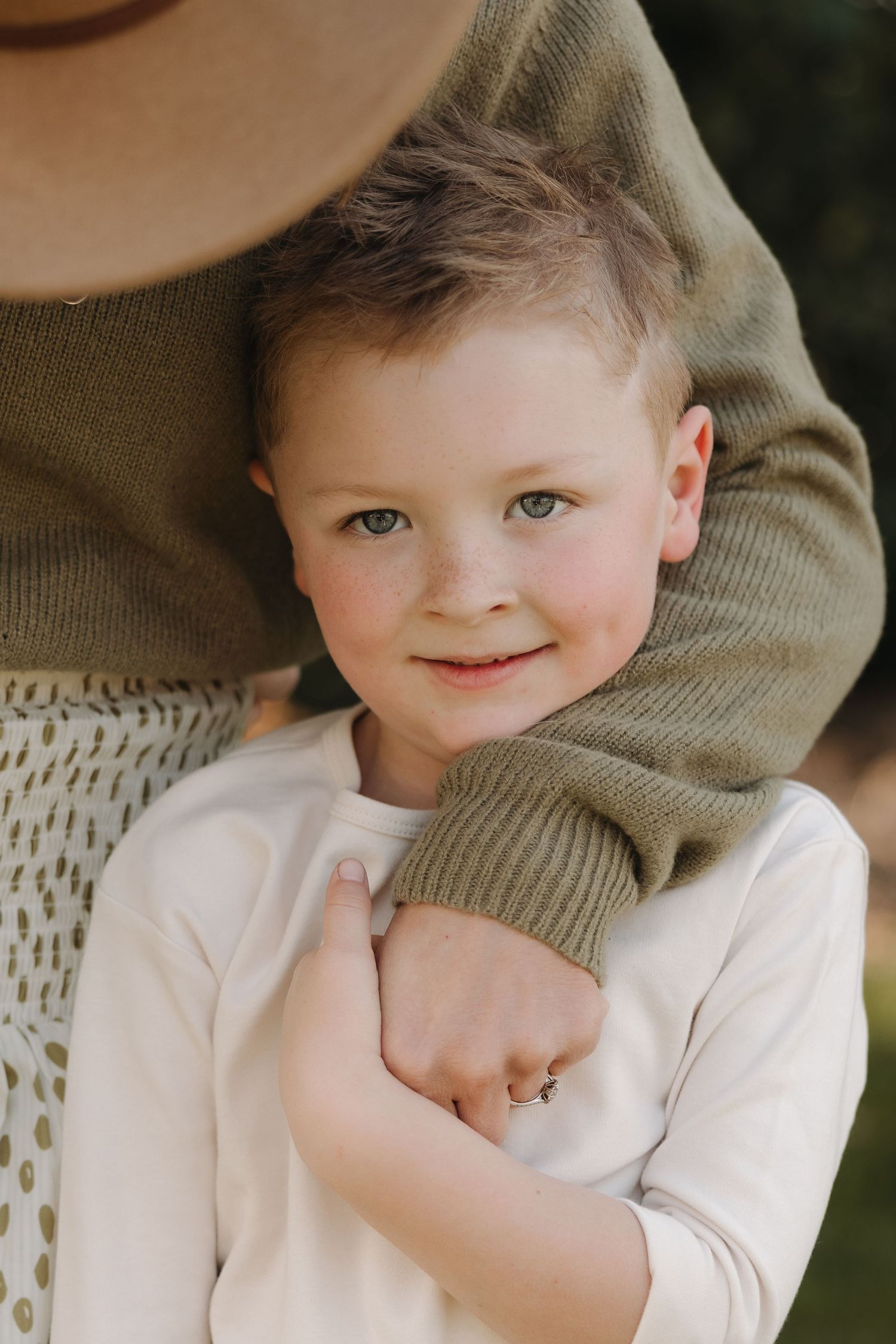 A child in a white shirt and olive sweater sits comfortably while looking at the camera.
