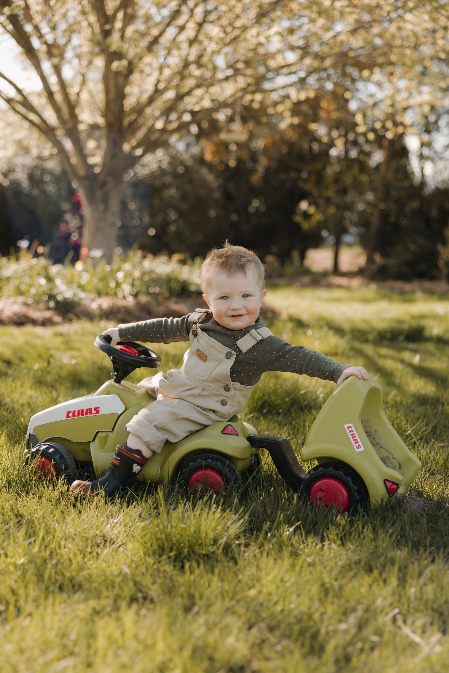 A toddler plays with a green Claas toy tractor while laying in grass on a sunny day.
