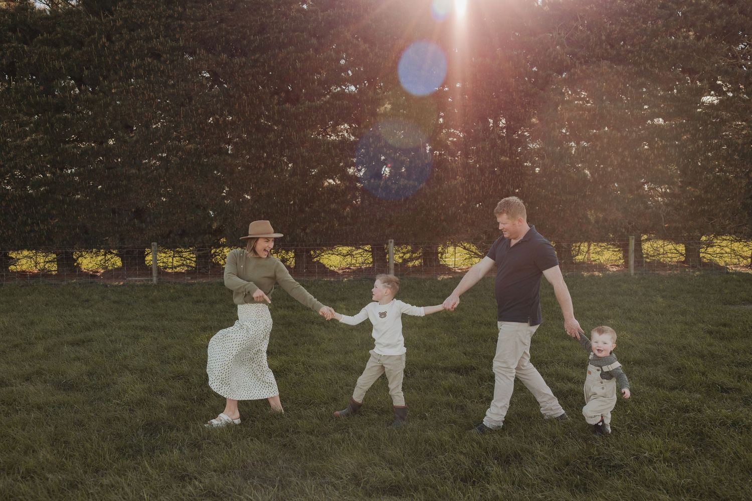 A family holds hands in a circle on a grassy field at sunset with sunflare in the background.