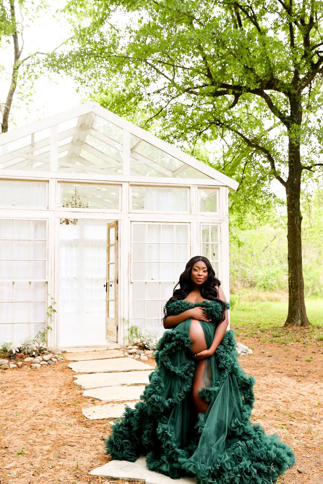 Elegant maternity photoshoot featuring a flowing emerald green gown in front of a white greenhouse structure outdoors.