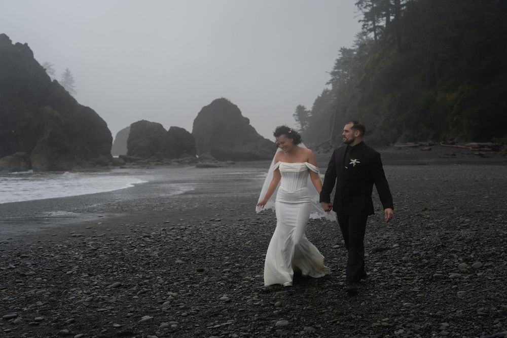 Couple walking hand in hand on a misty black sand beach with dramatic rock formations along the Pacific Northwest coast.