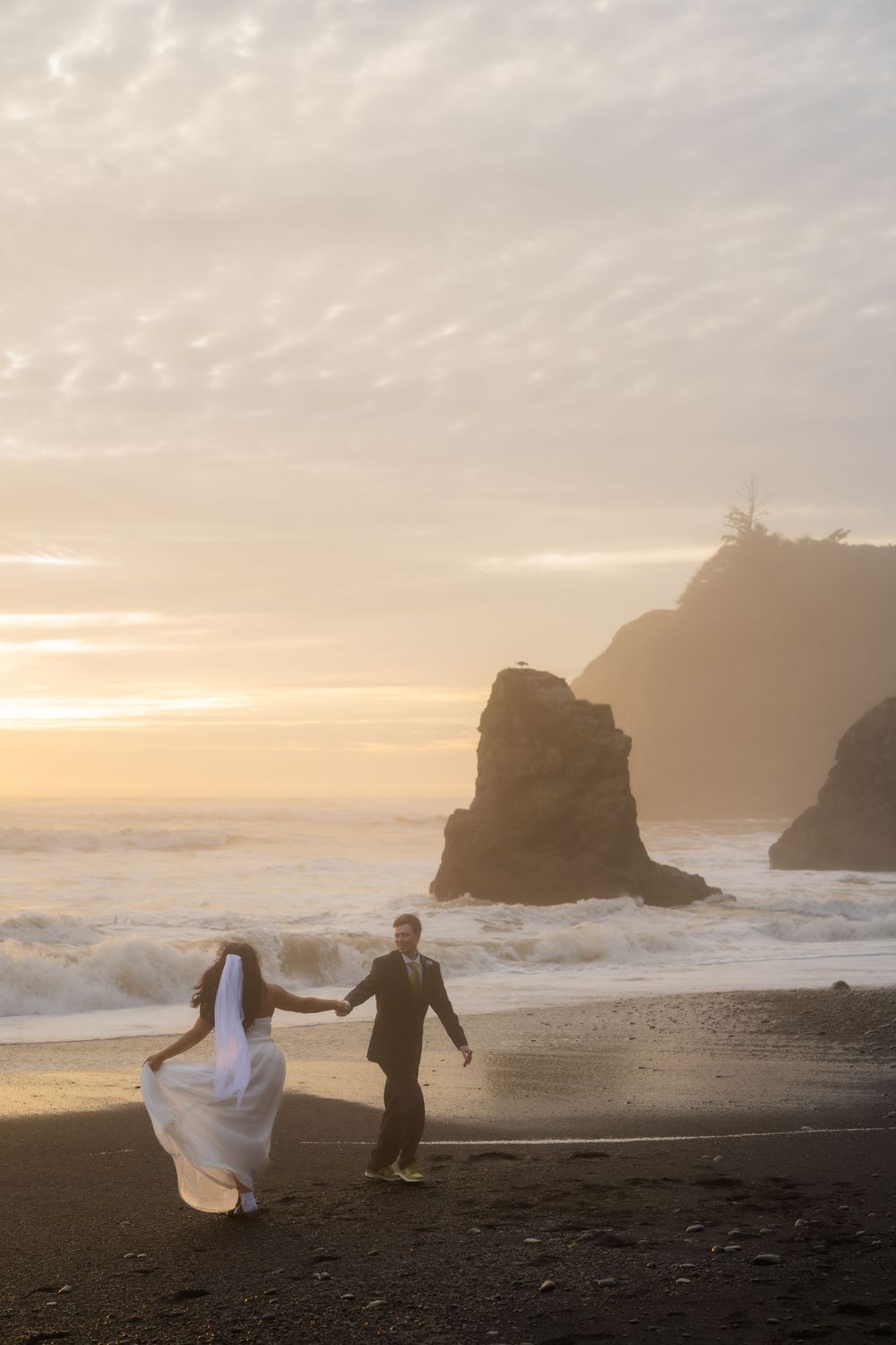 A couple dances on a misty beach at sunset with dramatic coastal cliffs and crashing waves in the background.