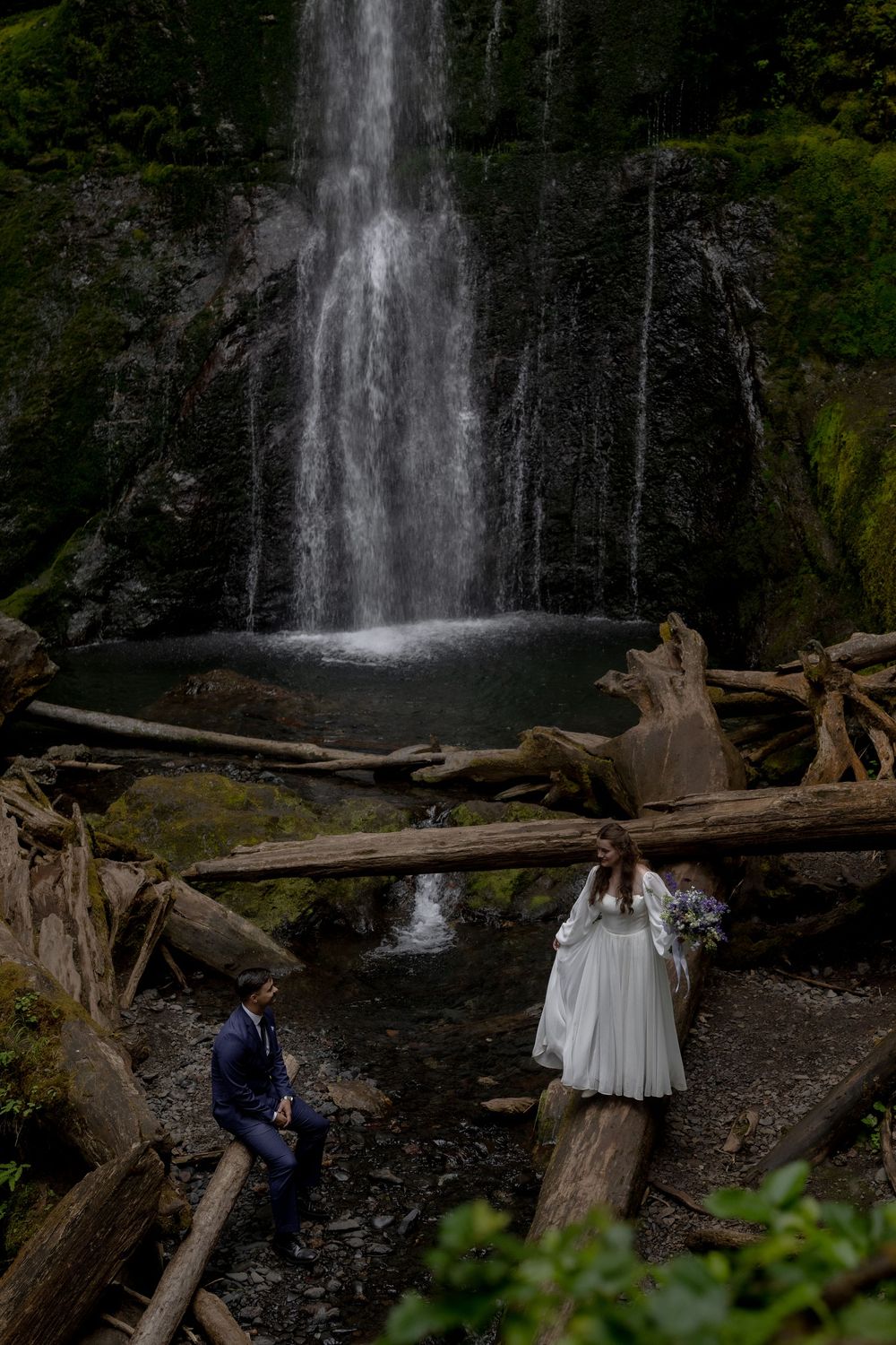 A couple shares an intimate moment in front of a majestic waterfall cascading down rocky cliffs surrounded by fallen logs.