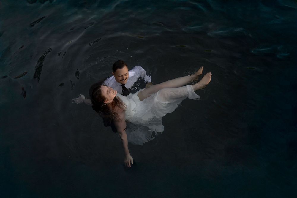 Couple in wedding attire lies romantically on wooden dock over calm lake at dusk.