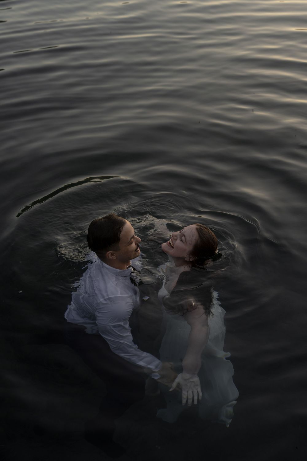 Two figures embrace while floating in dark rippling water at dusk creating a romantic mood.