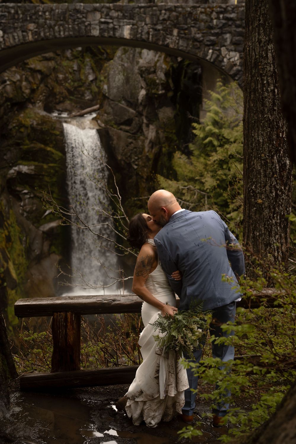 Two people share an intimate moment while overlooking a scenic waterfall in a lush forest setting.