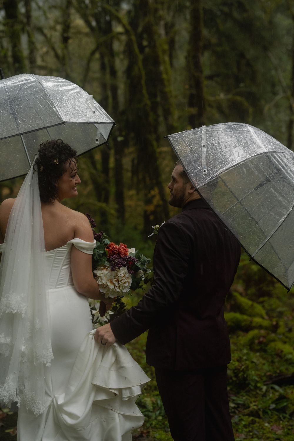A couple shares an intimate moment under clear umbrellas in a misty forest during their rainy wedding day.