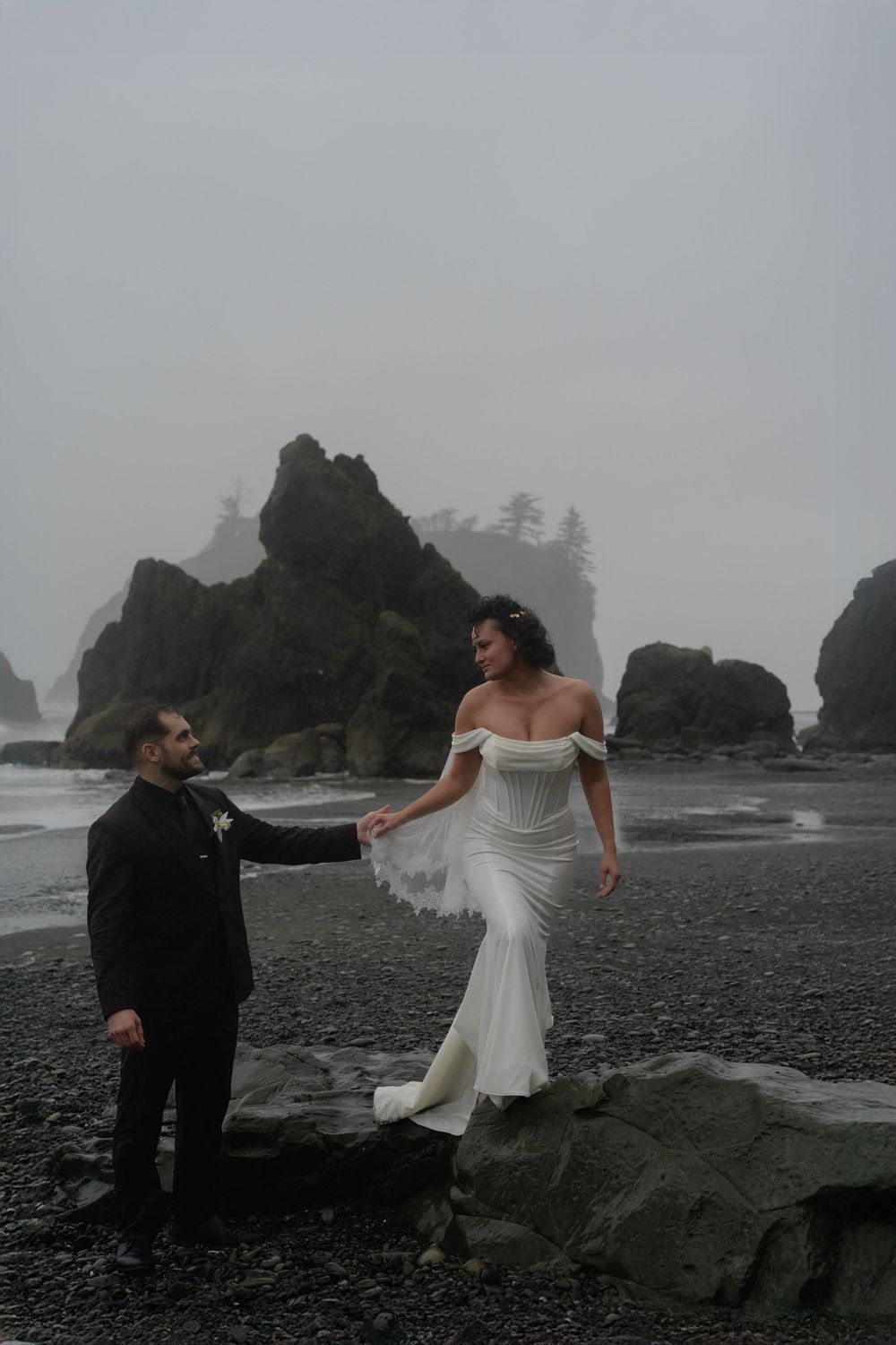 A couple in wedding attire poses dramatically on a misty rocky beach with dark cliffs and evergreen trees in the background.