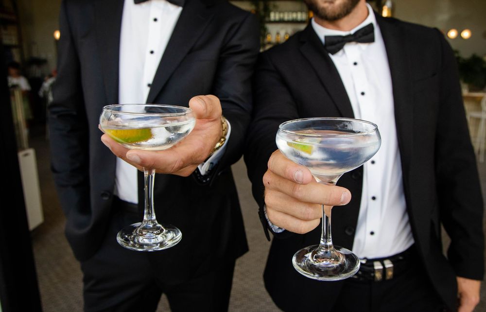 Two people in tuxedos holding elegant cocktail glasses with garnished martinis at a formal event.