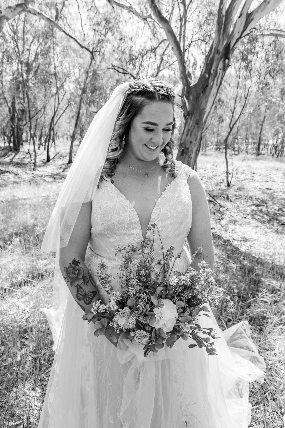 A bride in a lace wedding dress holds a rustic bouquet in a woodland setting.