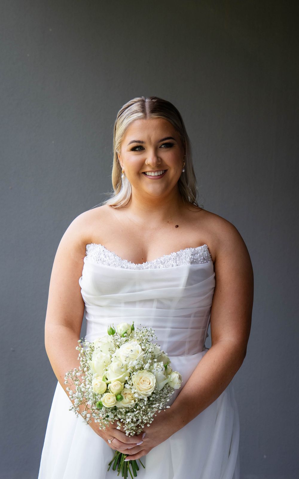 A bride in a white strapless gown holds a delicate bouquet of white roses and baby's breath against a gray backdrop.