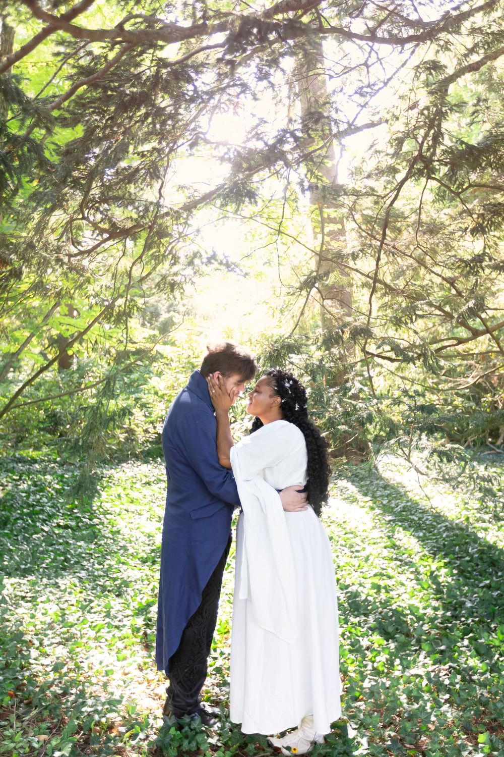 A couple in wedding attire share a romantic moment in a sunlit forest clearing surrounded by lush green trees.