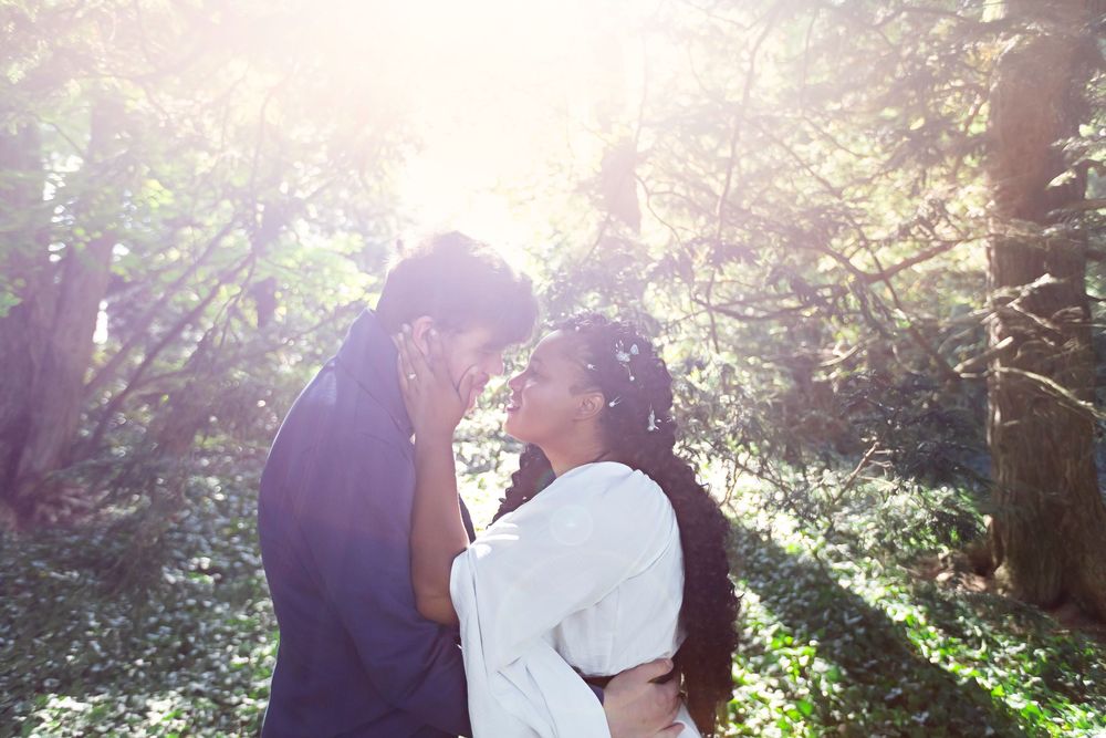 Couple sharing a romantic almost-kiss in a sunlit forest, surrounded by lush green trees and soft natural light.