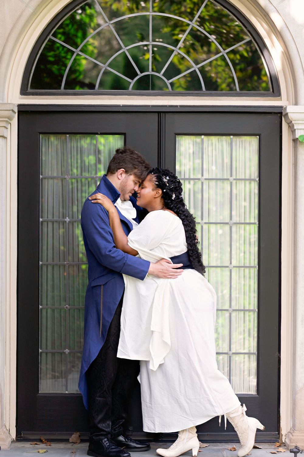 Interracial couple in elegant wedding attire embracing romantically in front of ornate black double doors.