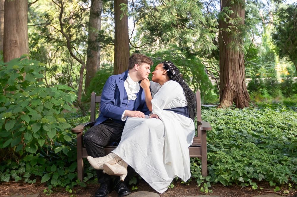 Couple in period costumes sharing a romantic moment on a wooden bench surrounded by lush green trees.