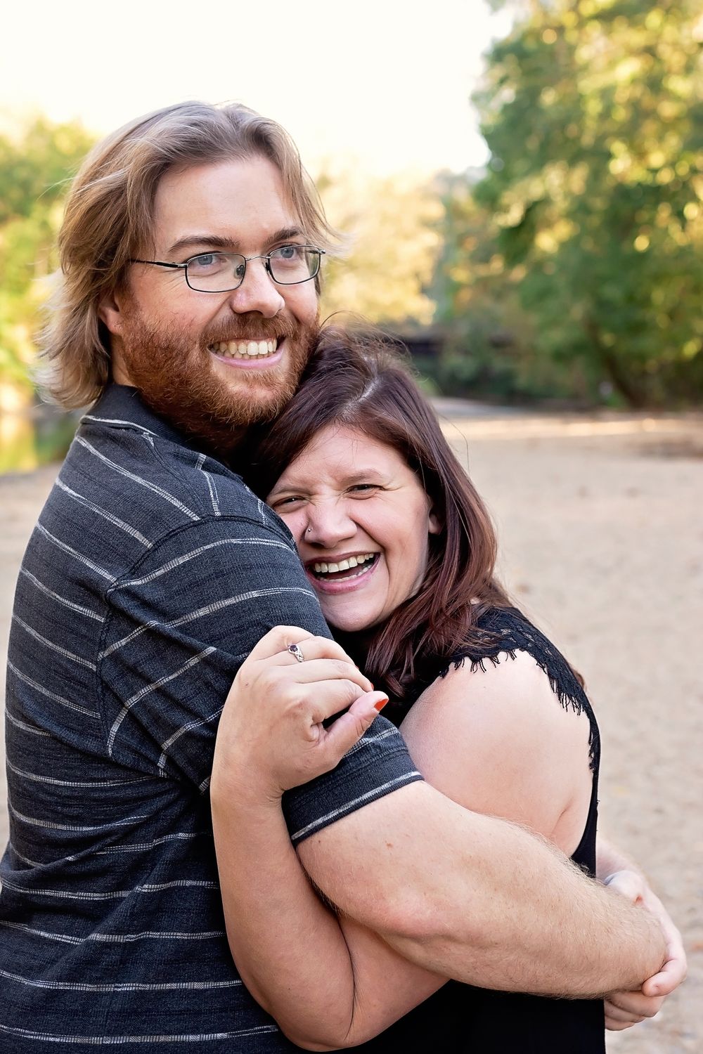 A smiling couple embraces outdoors, the bearded man with glasses hugging a laughing woman in a sandy park setting.