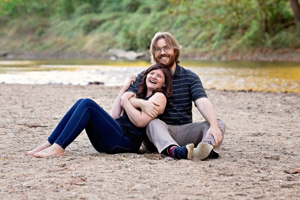Couple sitting on sandy riverbank laughing, man with long hair in striped shirt, woman in jeans leaning against him.