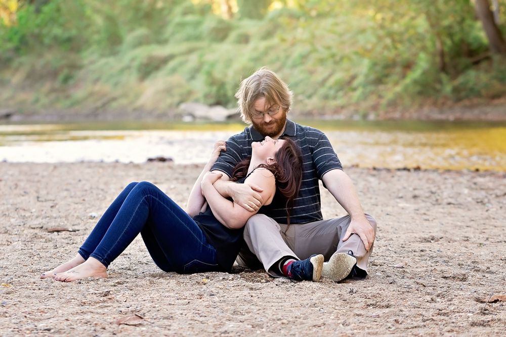 Couple sitting together on sandy riverbank, man with blonde hair embracing woman lying across his lap outdoors.