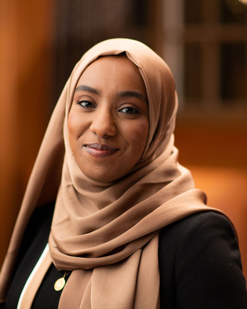Professional portrait of someone wearing a beige hijab and black blazer against a warm-toned background.