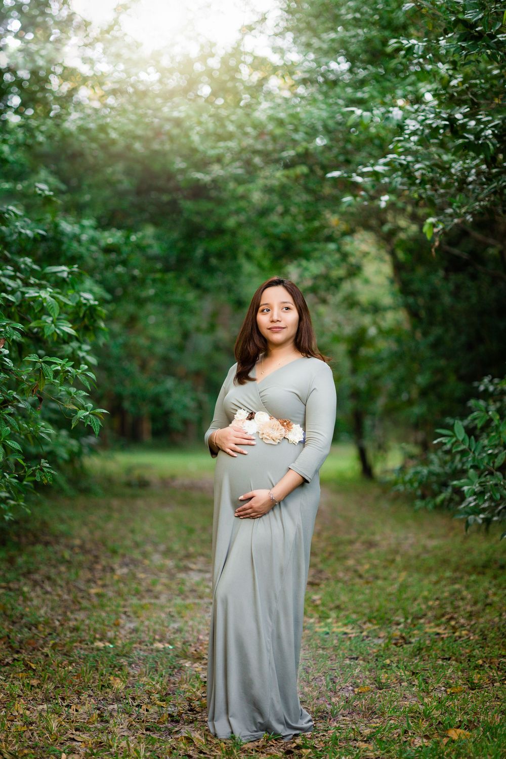 Pregnant woman in sage green maxi dress with floral sash stands on lush green garden path.