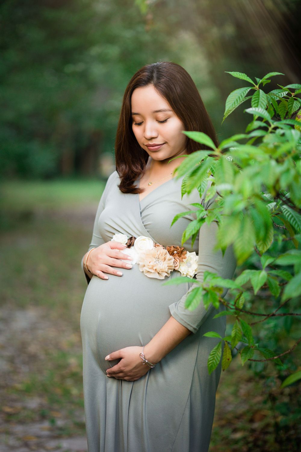 Pregnant woman in sage green wrap dress cradles baby bump beside green foliage in park setting.