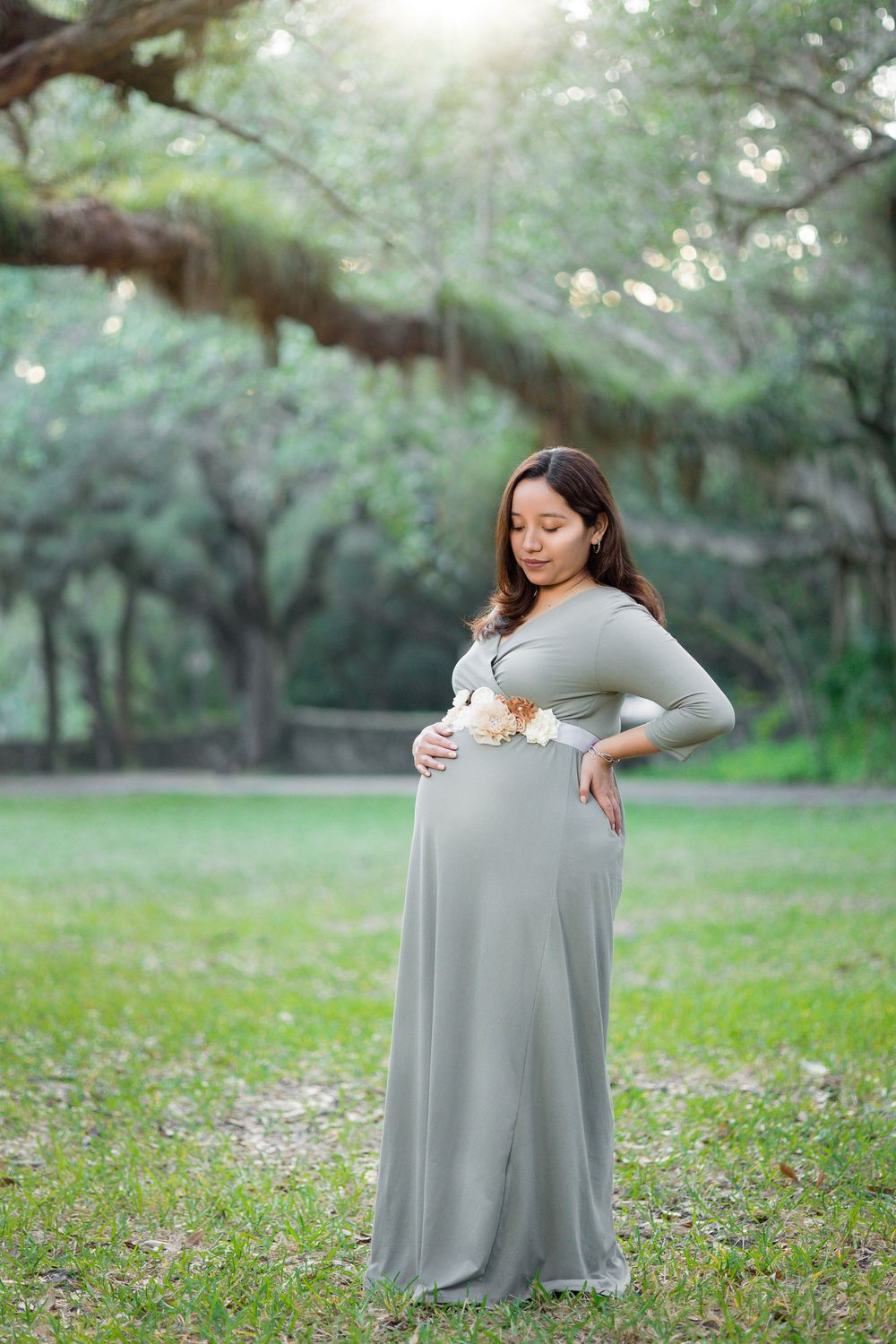 Pregnant woman in sage green maxi dress holds baby bump while standing under oak trees at sunset.