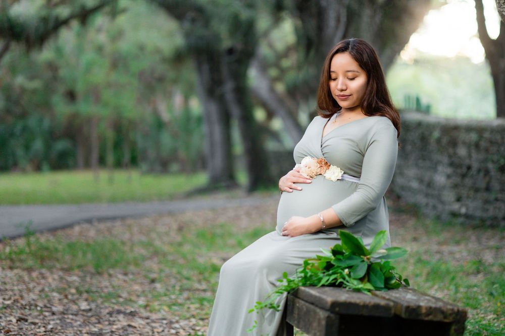 Pregnant woman in sage green maxi dress sits on rustic bench cradling bump in serene park.