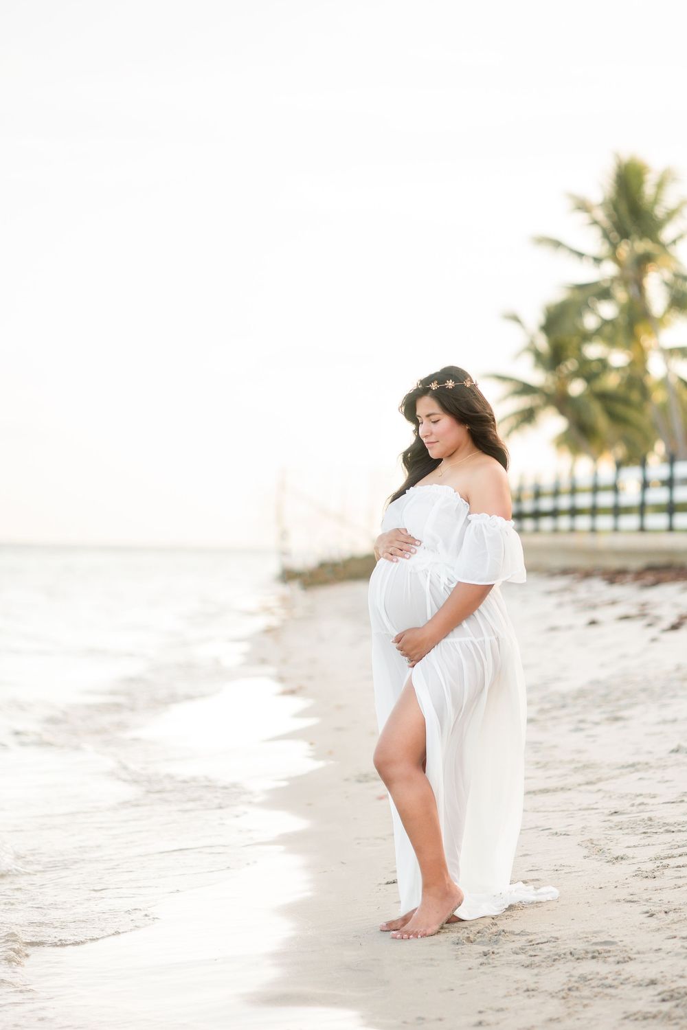 A glowing pregnant woman in a white maternity gown gently cradles her baby bump on a tranquil sandy beach.