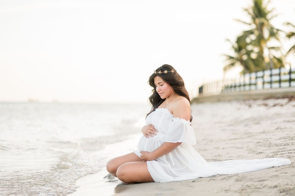A pregnant woman in a white off-shoulder dress sits on the beach, tenderly cradling her baby bump at sunset.