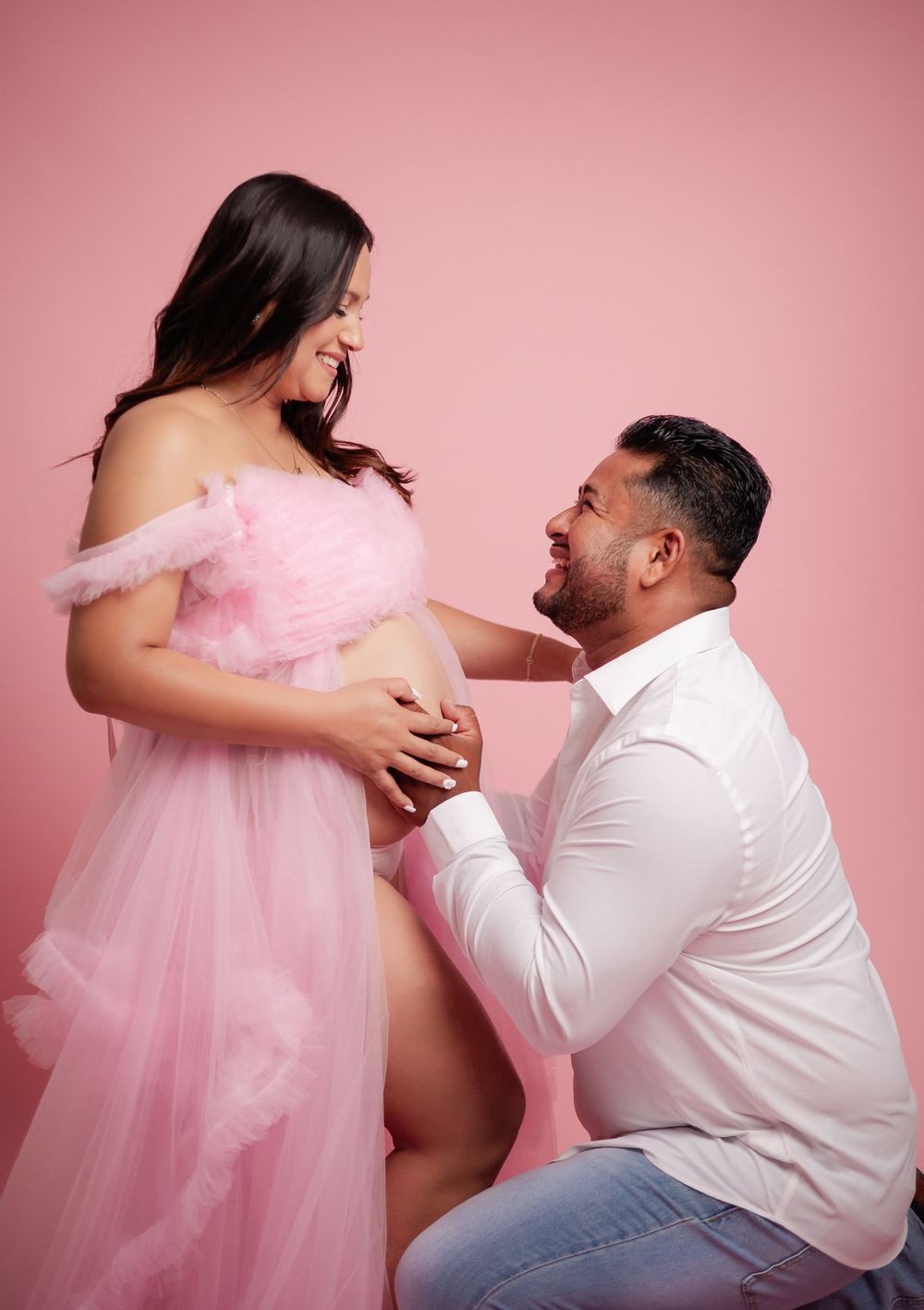 Couple laughing together during maternity shoot, pregnant woman in pink tulle gown on pink backdrop.