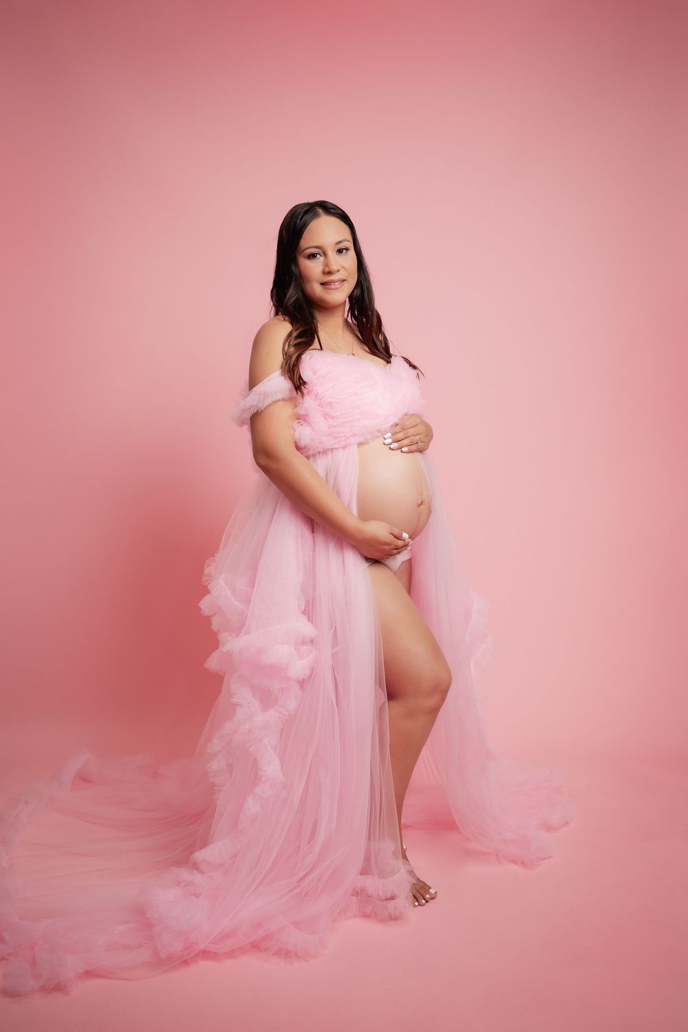 Pregnant woman in flowing pink tulle maternity gown cradling baby bump against pink studio background.