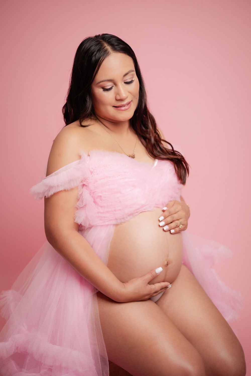 Pregnant woman in pink off-shoulder tulle gown cradles bare baby bump against soft pink background.